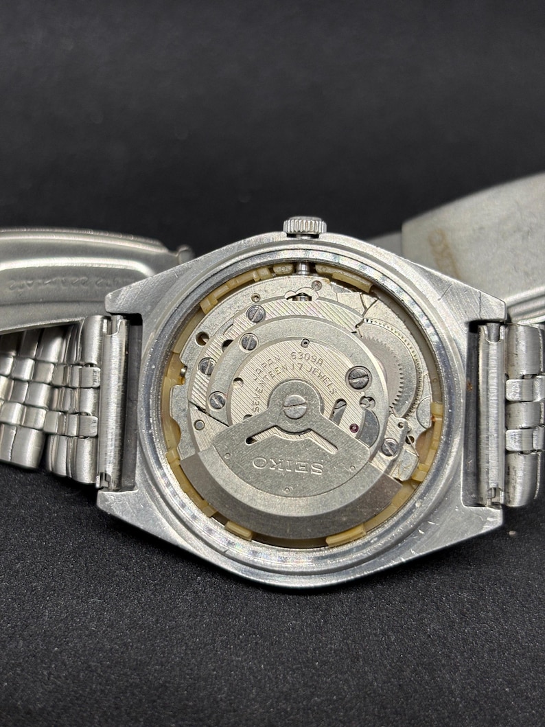 Peut inclure: Une montre-bracelet Seiko argent&eacute;e avec le bo&icirc;tier arri&egrave;re retir&eacute; pour r&eacute;v&eacute;ler le mouvement m&eacute;canique interne. Le m&eacute;canisme en m&eacute;tal porte les inscriptions 'SEIKO', '6309A' et 'SEVENTEEN 17 JEWELS' grav&eacute;es sur la masse oscillante.