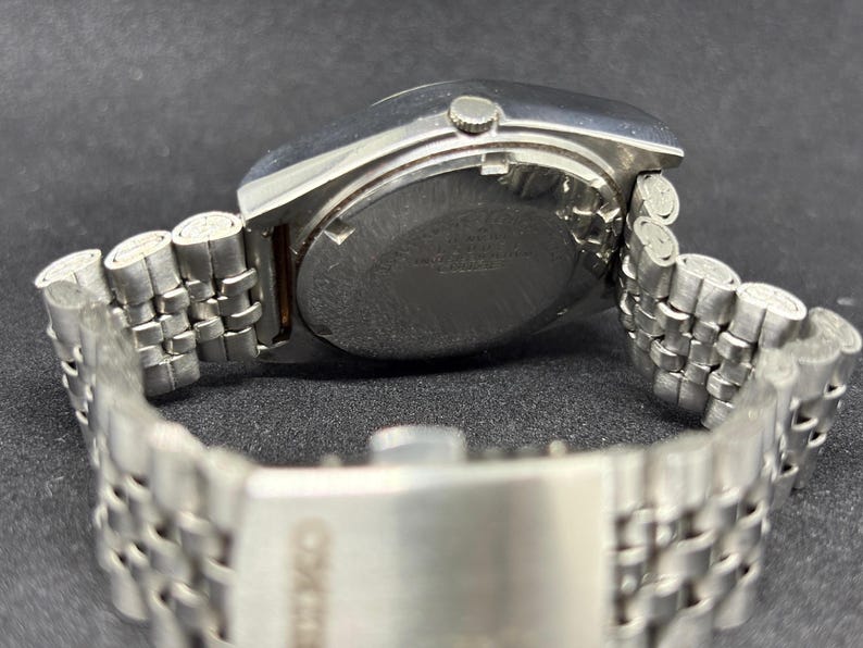 Peut inclure: Une montre-bracelet en acier inoxydable argent&eacute; avec un bo&icirc;tier rond et un bracelet en m&eacute;tal &agrave; maillons multiples. La montre comprend une petite couronne sur le c&ocirc;t&eacute; et un fermoir grav&eacute; du nom de la marque Seiko, montrant le dos du bo&icirc;tier.