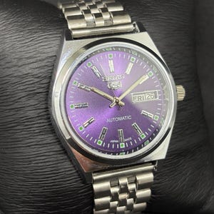 Peut inclure: Une montre automatique Seiko 5 en acier inoxydable dot&eacute;e d'un cadran violet &eacute;clatant, d'index argent&eacute;s avec des accents verts lumineux, d'un guichet jour-date &agrave; trois heures et d'un bracelet &agrave; maillons en m&eacute;tal argent&eacute; assorti.