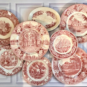 May include: A collection of eight ceramic plates and one oval platter featuring red transferware patterns. Each piece displays detailed historical scenes, architectural landmarks, or nautical imagery surrounded by intricate floral borders.