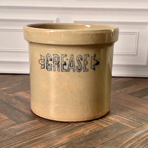 May include: A cylindrical tan stoneware crock featuring a wide rim and a flat base. The word GREASE is stamped in blue capital letters on the front, flanked by decorative arrow-like symbols on both sides of the text, creating a functional container.
