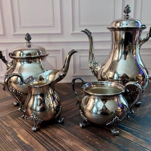 May include: A four-piece silver-plated tea service set featuring a tall coffee pot, a shorter teapot, a creamer, and a sugar bowl. Each piece has a polished metallic finish, ornate curved handles, decorative floral feet, and matching finials.