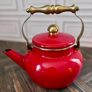 Op de afbeelding: Een felrode geëmailleerde theeketel met een handvat van gepolijst messing en een bijpassende messing knop op het deksel. De ketel heeft een ronde vorm, een korte tuit en een metalen messing rand rondom de opening waar het deksel op rust.