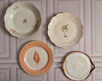 May include: Four decorative plates: one white with pink floral accents, one white with a green palm tree, one with a peach-colored border and a central seashell illustration, and one iridescent white plate with gold-toned handles and a gold rim.
