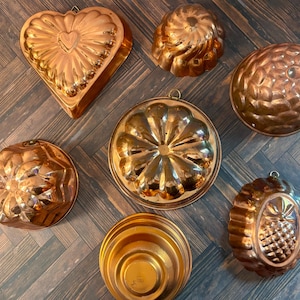 May include: A collection of seven copper-colored metal molds in various shapes, including a heart, a pineapple, and several fluted circular designs. Each mold features a small metal loop at the top edge for hanging, with a shiny, reflective finish.
