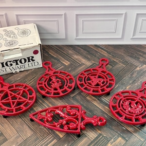 Può includere: Un set di cinque sottopentole in ghisa rossa con vari disegni traforati, tra cui una stella, una figura, un triscele, un motivo floreale e volute, accompagnati da una scatola di cartone bianca con l'etichetta VIGTOR CAST WARE LTD.