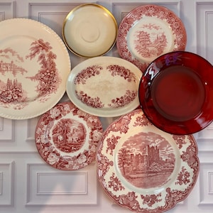 May include: A collection of six ceramic and glass plates and dishes. Four pieces feature cream backgrounds with red transferware landscape and floral patterns. One plate is solid red glass, and one small saucer has a gold rim.