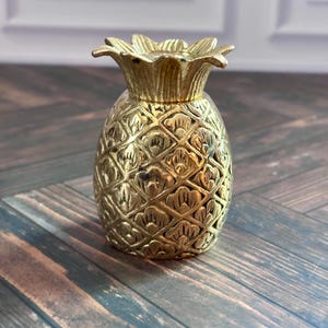 Può includere: Una piccola statuetta a forma di ananas in metallo color oro con un corpo testurizzato a motivo di rombi e una corona di foglie dettagliata. L'oggetto presenta una finitura metallica con riflessi e ombre che ne evidenziano la forma.