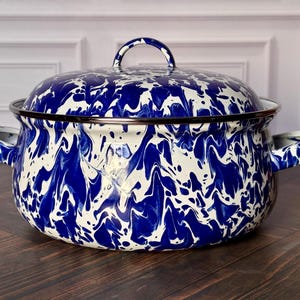 May include: A round, deep cooking pot with a matching lid, featuring a blue and white marbled enamel finish. The pot has two side handles and a curved lid handle, with a dark rim around the top edge of the base and the bottom edge of the lid.