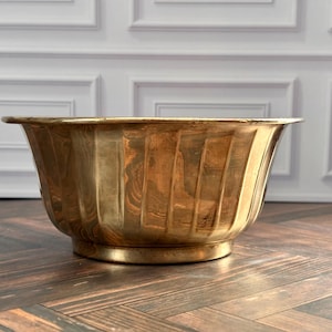 Op de afbeelding: Een grote, ronde koperen kom met een gepolijste metalen afwerking en een reeks verticale groeven langs de buitenwanden. De kom heeft een brede, uitlopende rand en een stevige, ronde voet, met een warm, goudkleurig oppervlak.