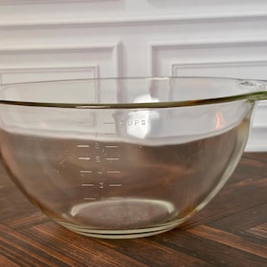 May include: A clear glass mixing bowl with a pouring spout and a handle. The exterior features etched measurement markings in cups, ranging from 2 to 8 cups, with the word CUPS printed clearly above the scale on the side of the bowl.