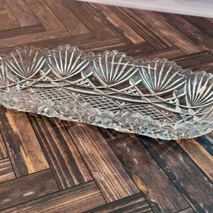 Può includere: Un vassoio da portata allungato in vetro trasparente con bordo smerlato e un motivo pressato dettagliato. Il design presenta motivi a ventaglio ripetuti lungo il bordo e una trama a rombi incrociati che copre il centro del piatto.