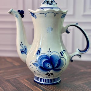 May include: A white ceramic teapot featuring hand-painted blue floral motifs and scrolling patterns. The teapot has a curved spout, a matching lid with a decorative finial, a sturdy handle, and a base decorated with vertical blue stripes.