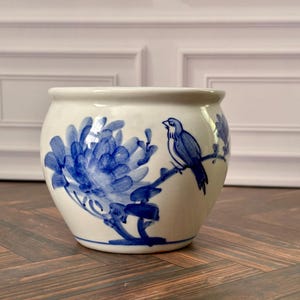 Può includere: Un vaso per piante in ceramica bianca con un motivo floreale blu dipinto a mano e un uccellino appollaiato su un ramo. Il contenitore arrotondato ha una finitura liscia e un'ampia apertura, con una classica colorazione blu e bianca.