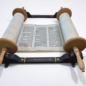 May include: A Torah scroll featuring Hebrew text on parchment, mounted on two light-colored wooden rollers. The scroll is supported by a black wooden stand with gold-colored Hebrew lettering and decorative accents on the front crossbar.