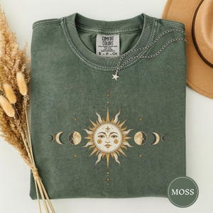 May include: A moss green crewneck t-shirt featuring a central graphic of a sun with a human face surrounded by moon phases and small stars. The shirt has a ribbed collar and a Comfort Colors label. A silver star pendant necklace rests on the fabric.