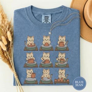 May include: A blue t-shirt featuring a graphic of nine light brown cats working with clay on pottery wheels. The shirt has a crew neck and a Comfort Colors label at the inner collar. The graphic depicts various stages of the pottery process.