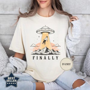 May include: An ivory t-shirt featuring a graphic of a UFO beaming up a person above a mountain range. The design includes a sunset, clouds, and the text "FINALLY" printed in black below the mountains. The shirt has a crew neckline and short sleeves.