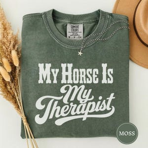 Op de afbeelding: Een mosgroen t-shirt met korte mouwen, een ronde hals en een witte grafische opdruk met de tekst My Horse Is My Therapist. Het shirt is voorzien van een zilverkleurige ketting met een kleine sterhanger bij de kraag.