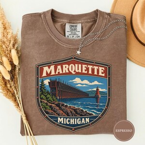 Può includere: Una t-shirt marrone a girocollo con una grafica del molo di carico del minerale e del faro di Marquette, Michigan. Il disegno è racchiuso in un bordo a forma di scudo con rivetti. La maglietta ha un'etichetta Comfort Colors e una collana a stella.