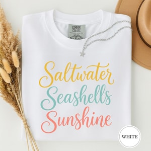 以下が含まれることがあります： 黄色、ティール、コーラルの筆記体で「Saltwater Seashells Sunshine」とプリントされた白い半袖Tシャツ。Tシャツの内側の襟元にはComfort Colorsのブランドラベルがあり、銀色の星のペンダントネックレスが添えられています。