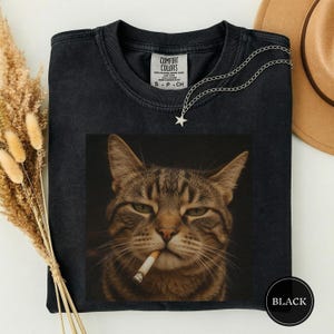 May include: A black crew neck t-shirt featuring a graphic print of a brown tabby cat with a cigarette in its mouth. The shirt has a Comfort Colors label at the inner neckline and is paired with a silver star pendant necklace on a thin chain.