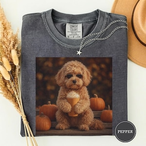 May include: A charcoal gray crew neck t-shirt featuring a graphic of a fluffy brown poodle wearing sunglasses and holding a cup of coffee. The poodle is surrounded by orange pumpkins against a blurred autumn background. The shirt has a Comfort Colors label.