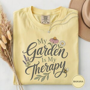 Op de afbeelding: Een lichtgeel t-shirt met korte mouwen en de donkergrijze tekst My Garden Is My Therapy. Het ontwerp bevat illustraties van een roze bloem, een wit madeliefje, lavendeltakjes, groene bladeren en een kleine bij. Het shirt heeft een S-label.