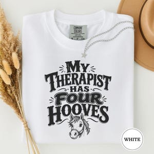 Op de afbeelding: Een wit t-shirt met ronde hals en de zwarte tekst "MY THERAPIST HAS FOUR HOOVES" boven een lijntekening van een paardenkop. Het shirt heeft een Comfort Colors-label bij de binnenhals en is gecombineerd met een zilveren sterrenketting.