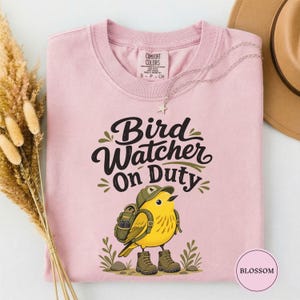 May include: A light pink t-shirt featuring a graphic of a yellow bird wearing a green cap and a backpack, standing in boots. The text reads Bird Watcher On Duty in black script. The shirt has a Comfort Colors label at the inner neckline.