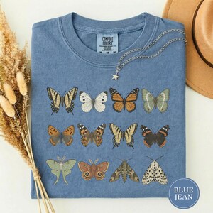 Puede incluir: Una camiseta azul con un estampado gráfico de once mariposas y polillas diferentes dispuestas en tres filas. La prenda tiene cuello redondo y una etiqueta de la marca Comfort Colors en el interior. Un collar con estrella plateada descansa sobre la tela.