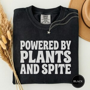 以下が含まれることがあります： 前面に白い文字で「POWERED BY PLANTS AND SPITE」とプリントされた黒の半袖Tシャツ。クルーネックで、襟の内側にはComfort Colorsのブランドラベルが付いています。シルバーの星型ペンダントネックレスがTシャツの上に置かれています。