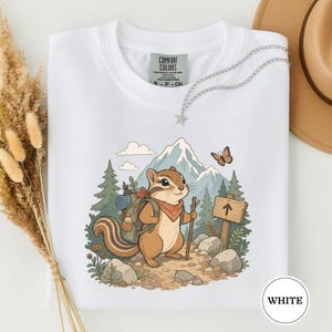 Op de afbeelding: Een wit T-shirt met ronde hals en een afbeelding van een chipmunk met een rugzak en bandana, die een wandelstok vasthoudt. De scène bevat dennenbomen, bergen, een houten bord met een pijl en een vlinder. Het label vermeldt Comfort Colors.