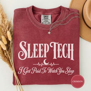 Könnte beinhalten: Ein karmesinrotes Kurzarm-T-Shirt mit Rundhalsausschnitt. Auf der Vorderseite befindet sich ein weißer Schriftzug "SLEEP TECH" über einer Grafik mit einem Halbmond und einer Herzschlaglinie, gefolgt vom Text "I Get Paid To Watch You Sleep".