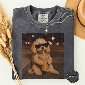 May include: A charcoal grey crewneck t-shirt featuring a graphic of a light brown dog wearing black sunglasses and holding a plastic cup with a straw. The shirt has a Comfort Colors label at the neck and a small star-shaped pendant necklace.