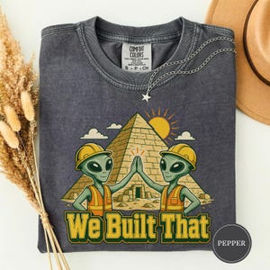 Könnte beinhalten: Ein anthrazitfarbenes T-Shirt mit Rundhalsausschnitt und einer Grafik von zwei grünen Außerirdischen, die gelbe Bauhelme und orangefarbene Warnwesten tragen und vor einer Pyramide und einer Sonne abklatschen. Der Text lautet We Built That in Gelb.