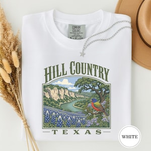 May include: A white t-shirt featuring a graphic print of a landscape with a river, cliffs, bluebonnet flowers, and a painted bunting bird. The text reads Hill Country Texas in a green serif font. A silver star necklace rests on the neckline.