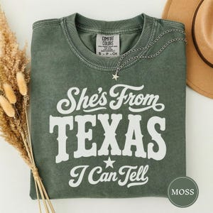 Könnte beinhalten: Ein moosgrünes Kurzarm-T-Shirt mit weißem Siebdrucktext She's From TEXAS I Can Tell und einer kleinen Sternengrafik. Das Shirt hat einen Rundhalsausschnitt und ein Comfort Colors Markenetikett an der Innenseite des Kragens.