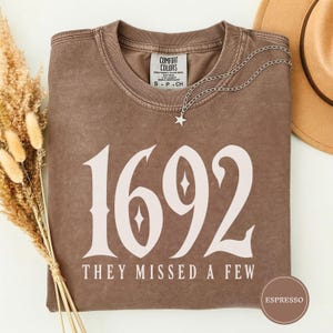 May include: A brown short-sleeved t-shirt with a vintage wash, featuring the large white text 1692 and the smaller text THEY MISSED A FEW below it. The shirt has a crew neckline and a Comfort Colors label. A silver star necklace rests on the collar.