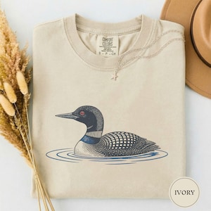 May include: An ivory-coloured t-shirt featuring a graphic illustration of a loon swimming in blue water. The bird has a black head, a red eye, and a patterned body. The shirt has a crew neckline and a label that reads COMFORT COLORS.