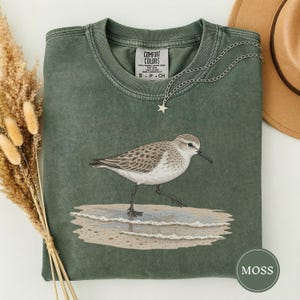 Puede incluir: Una camiseta de cuello redondo color verde musgo con una ilustración detallada de un correlimos caminando sobre una playa de arena con olas suaves. La prenda tiene un efecto de lavado vintage y una etiqueta Comfort Colors. Un collar de estrella plateado.
