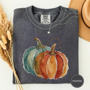 May include: A charcoal gray crew neck t-shirt with a watercolor-style graphic of two pumpkins, one teal and one orange, featuring a silver star necklace resting on the collar and a label reading Comfort Colors, with the color name Pepper.