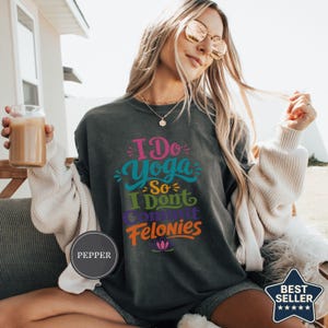 May include: A dark grey short-sleeve t-shirt featuring the colourful text I Do Yoga So I Dont Commit Felonies with a small lotus flower graphic at the bottom. The shirt has a crew neckline and a relaxed fit, showcasing a screen-printed design.