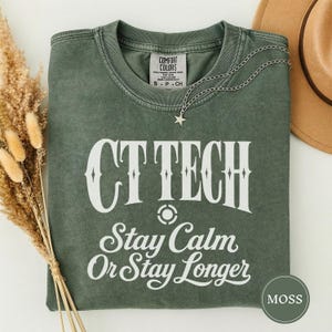 May include: A moss green crewneck t-shirt featuring the white text CT TECH and Stay Calm Or Stay Longer with a small star necklace resting on the collar. The shirt has a Comfort Colors label and a vintage-style wash finish on the fabric.