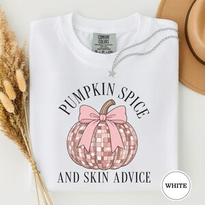 May include: A white short-sleeve t-shirt featuring a graphic of a pink disco ball pumpkin with a large pink bow. The text PUMPKIN SPICE AND SKIN ADVICE is printed in black around the pumpkin. The shirt has a Comfort Colors brand neck label.