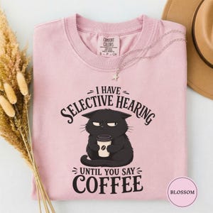 May include: A light pink t-shirt featuring a graphic of a grumpy black cat holding a coffee cup. The text reads I HAVE SELECTIVE HEARING UNTIL YOU SAY COFFEE. The shirt has a crew neckline and a Comfort Colors brand label printed on the inside neck.