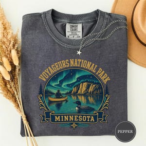 Puede incluir: Una camiseta de cuello redondo gris carbón con un gráfico del Parque Nacional Voyageurs en Minnesota. El diseño muestra a una persona en canoa en un lago con un colimbo, acantilados y auroras boreales. El texto dice Voyageurs National Park Minnesota.