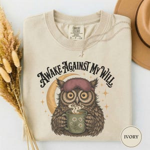 May include: An ivory t-shirt featuring a graphic of an owl wearing a sleep mask and holding a mug with a floral design. The text "AWAKE AGAINST MY WILL" is printed in a dark arched font above the owl, accompanied by a crescent moon and stars.