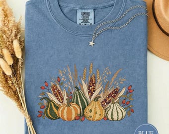Puede incluir: Una camiseta azul de manga corta con un estampado gráfico de varias calabazas, maíz y tallos de trigo. La prenda incluye una etiqueta Comfort Colors en el cuello. Una cadena plateada con un dije de estrella descansa sobre la tela.