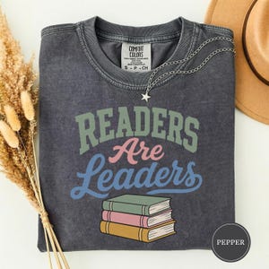 Könnte beinhalten: Ein anthrazitfarbenes Kurzarm-T-Shirt mit Vintage-Waschung. Die Grafik zeigt den Text "Readers Are Leaders" in Salbeigrün, Rosa und Blau sowie eine Illustration von drei gestapelten Büchern in Salbeigrün, Rosa und Gelb unter dem Text.